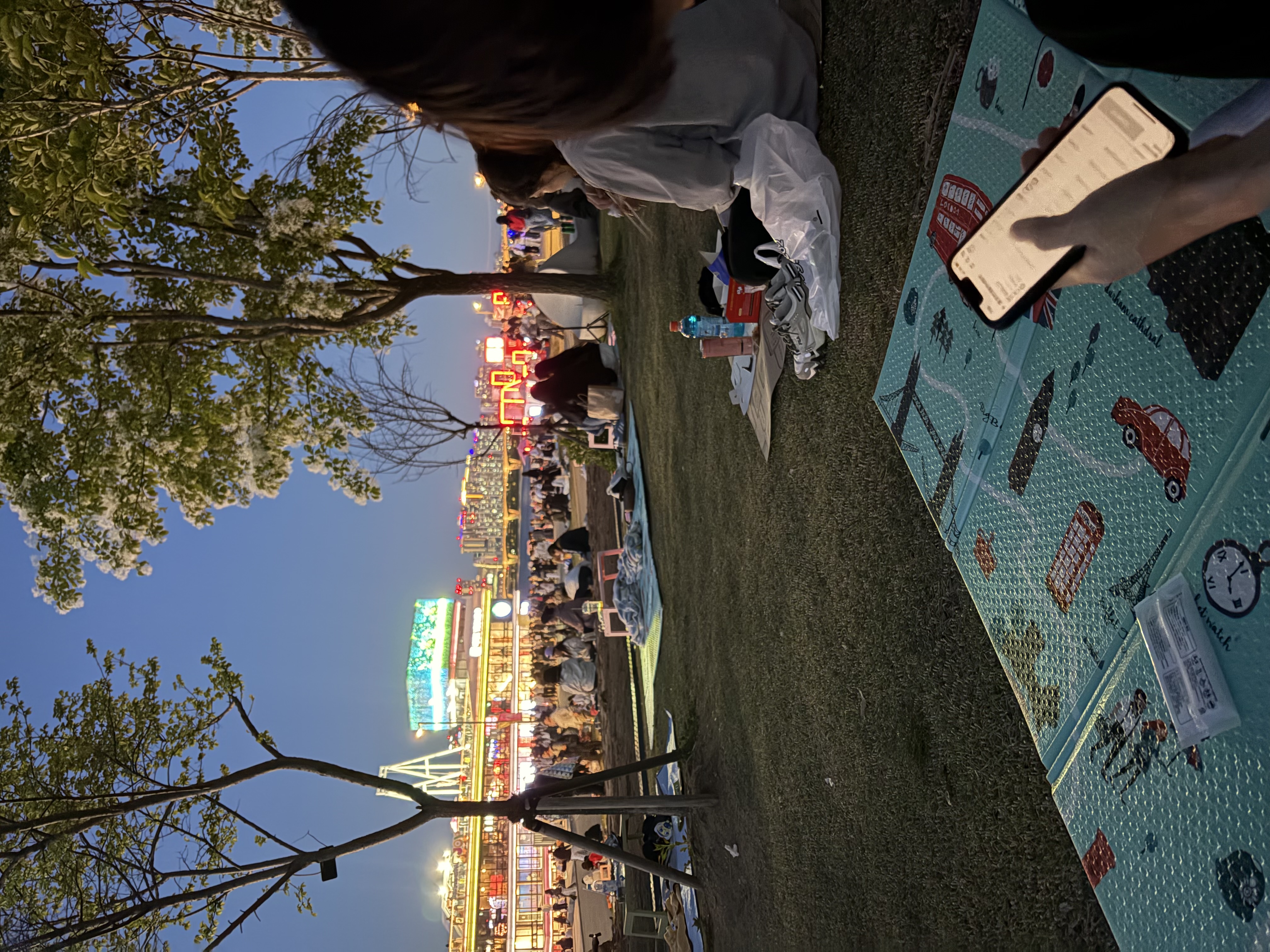 A lively evening scene by the river in Korea, with people sitting on the grass, city lights glowing in the background, and a relaxed, social atmosphere.
