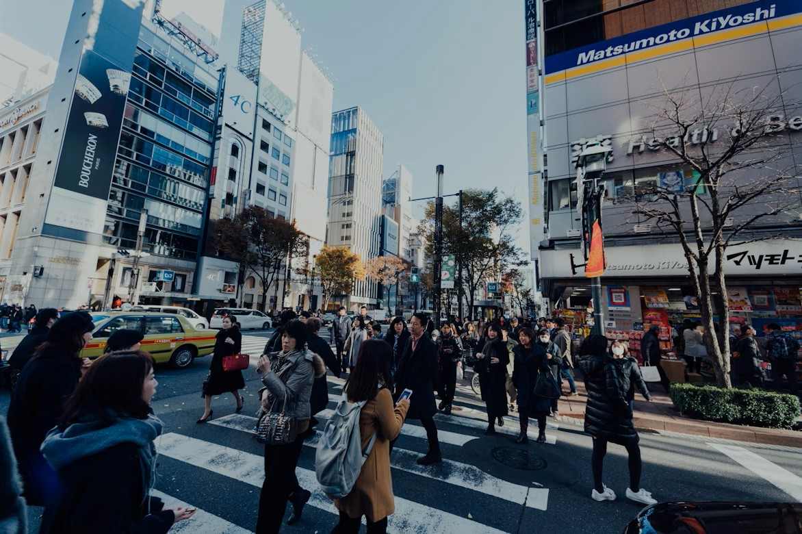 A busy street in Japan with many people crossing the road between tall buildings on a bright day.