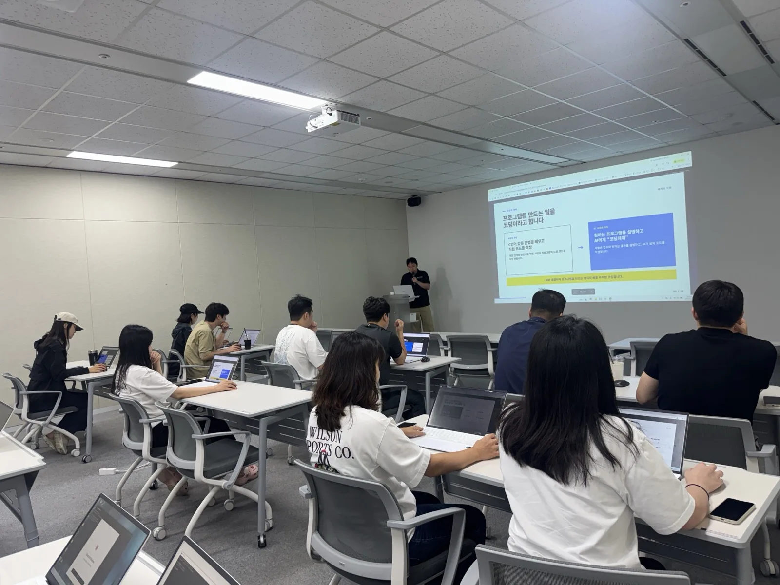 AI 응용 과정, 프로그램을 만드는 일을 코딩이라고 합니다 슬라이드 앞에서 강의하는 모습과 윌슨 티셔츠를 입은 수강생들